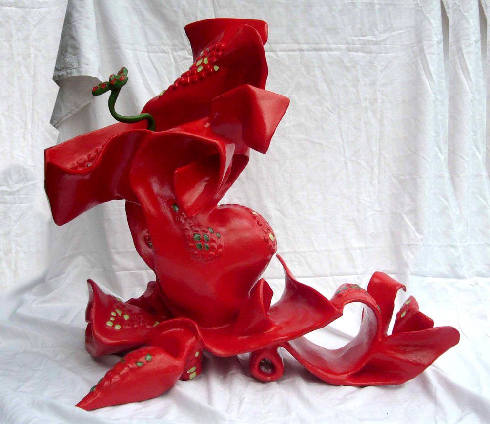 Abstrakte Plastik aus Pappmasché der Künstlerin Benita von Wendt. Titel: Daisy. Die abstrakte Skulptur ist rot mit hellgrünen Mosaiksteinen, anstatt eines Kopfes kommt aus ihrer Mitte heraus eine Blume. Die Plastik erinnert etwas an Daisy Duck, sie zieht eine Schleppe hinter sich her und hat übergroße Pömps an. Seitenansicht links.