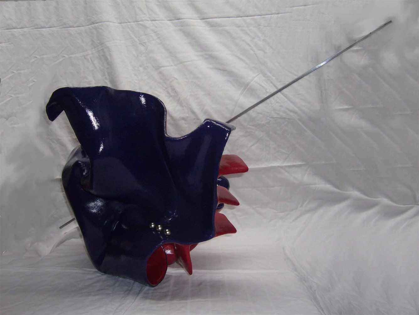 Abstrakte Plastik aus Pappmasché der Künstlerin Benita von Wendt. Titel: Don Quichotte. Die abstrakte Skulptur ist aussen Blau, innen rot, rechts sind Metallknöpfe befestigt, eine dünne Metallstange an der Skulptur erinnert an eine Lanze. Seitenansicht.