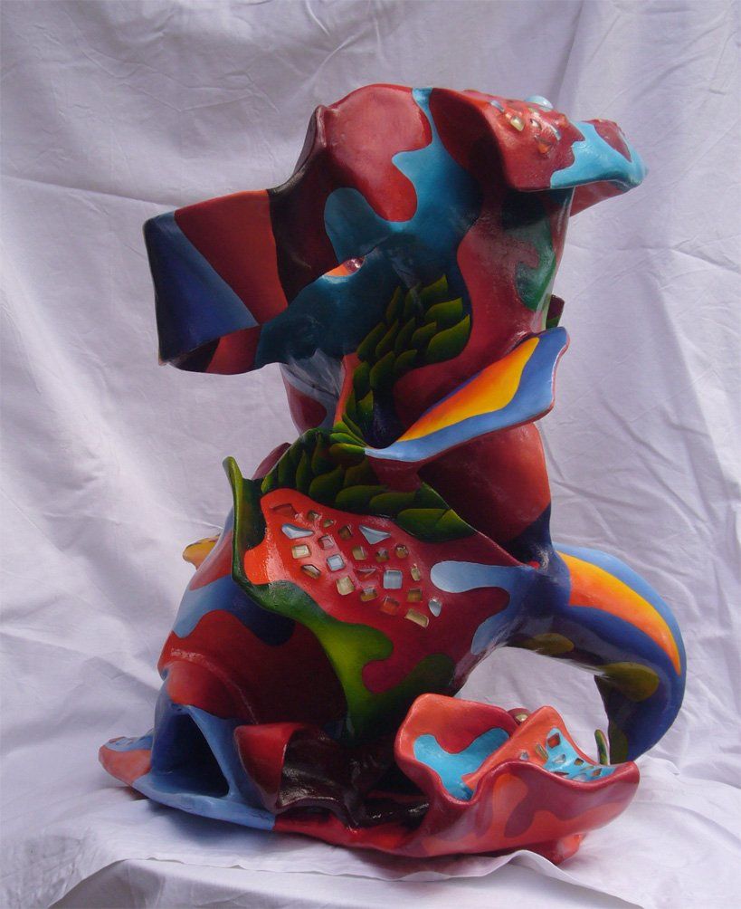 Abstrakte Plastik aus Pappmasché der Künstlerin Benita von Wendt. Titel: Elvis. Die abstrakte lebensfroh wirkende Skulptur ist sehr bunt mit Ornamenten die an Blätterwerk erinnern, sie scheint in einen Mantel mit sehr großem Kragen gehüllt. Im Innenraum des Kragens befindet sich eine Schublade, darauf sind mit Silberdraht zwei Augäpfel und ein Mund montiert. Seitenansicht links.
