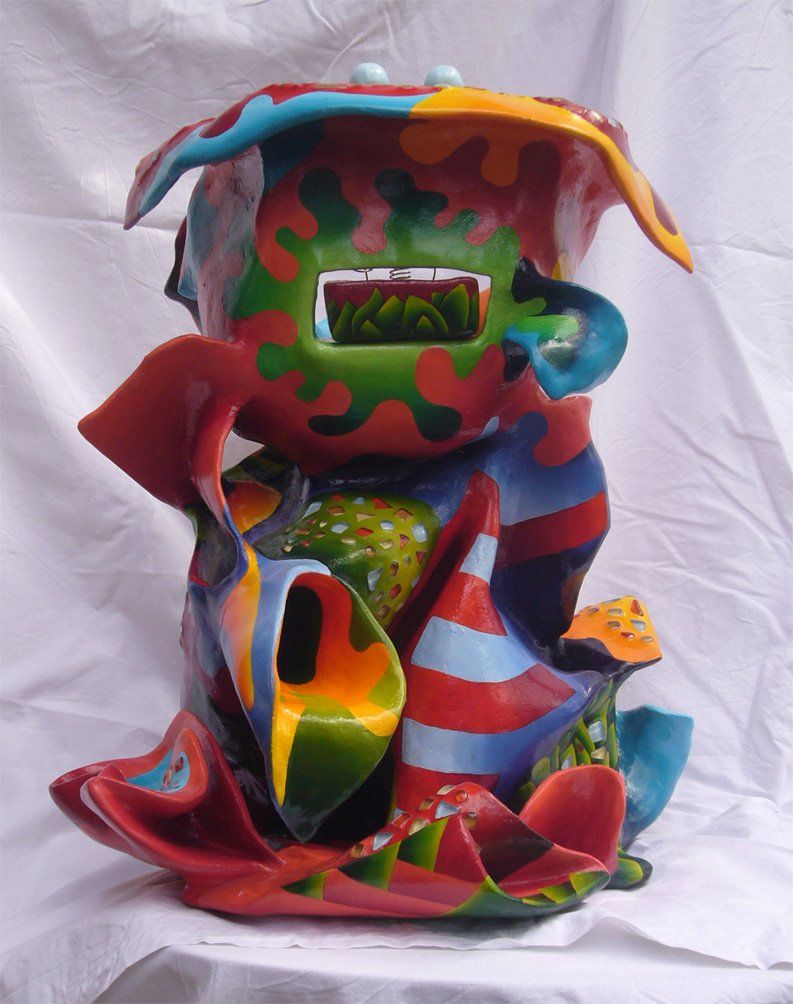 Abstrakte Plastik aus Pappmasché der Künstlerin Benita von Wendt. Titel: Elvis. Die abstrakte lebensfroh wirkende Skulptur ist sehr bunt mit Ornamenten die an Blätterwerk erinnern, sie scheint in einen Mantel mit sehr großem Kragen gehüllt. Im Innenraum des Kragens befindet sich eine Schublade, darauf sind mit Silberdraht zwei Augäpfel und ein Mund montiert. Rückansicht.