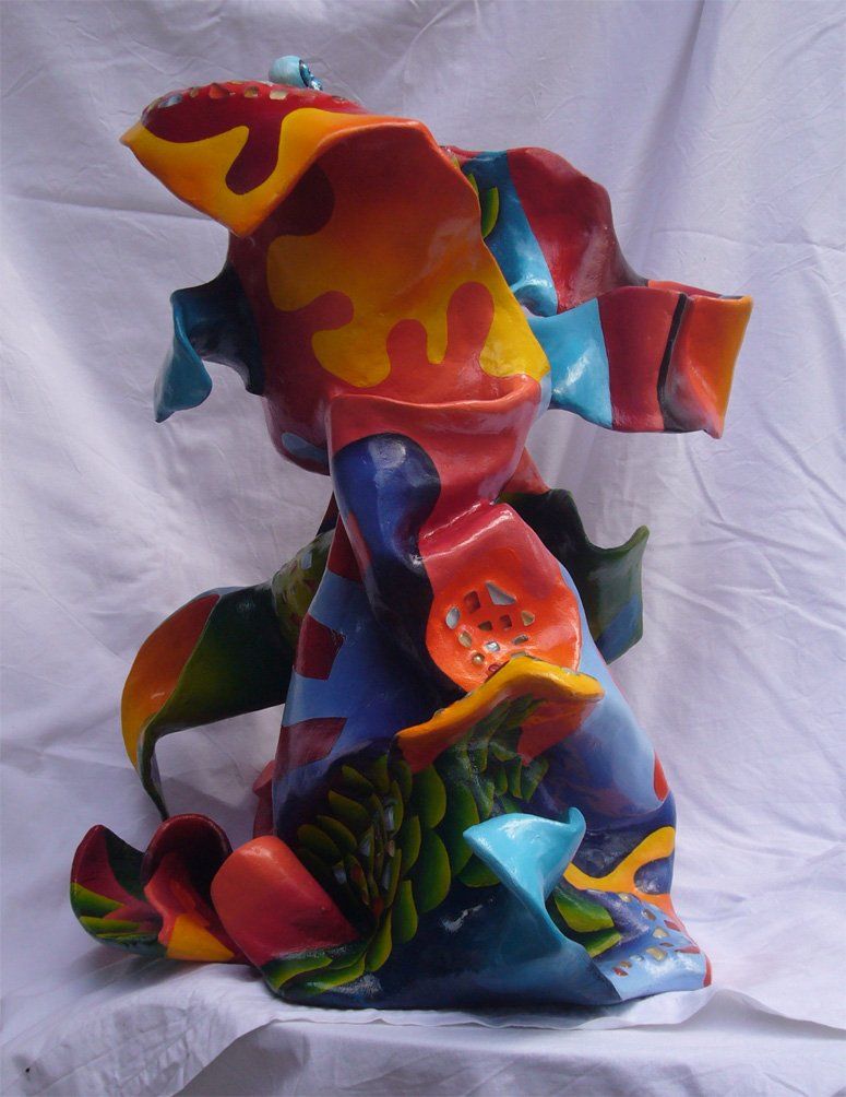 Abstrakte Plastik aus Pappmasché der Künstlerin Benita von Wendt. Titel: Elvis. Die abstrakte lebensfroh wirkende Skulptur ist sehr bunt mit Ornamenten die an Blätterwerk erinnern, sie scheint in einen Mantel mit sehr großem Kragen gehüllt. Im Innenraum des Kragens befindet sich eine Schublade, darauf sind mit Silberdraht zwei Augäpfel und ein Mund montiert. Seitenansicht rechts.