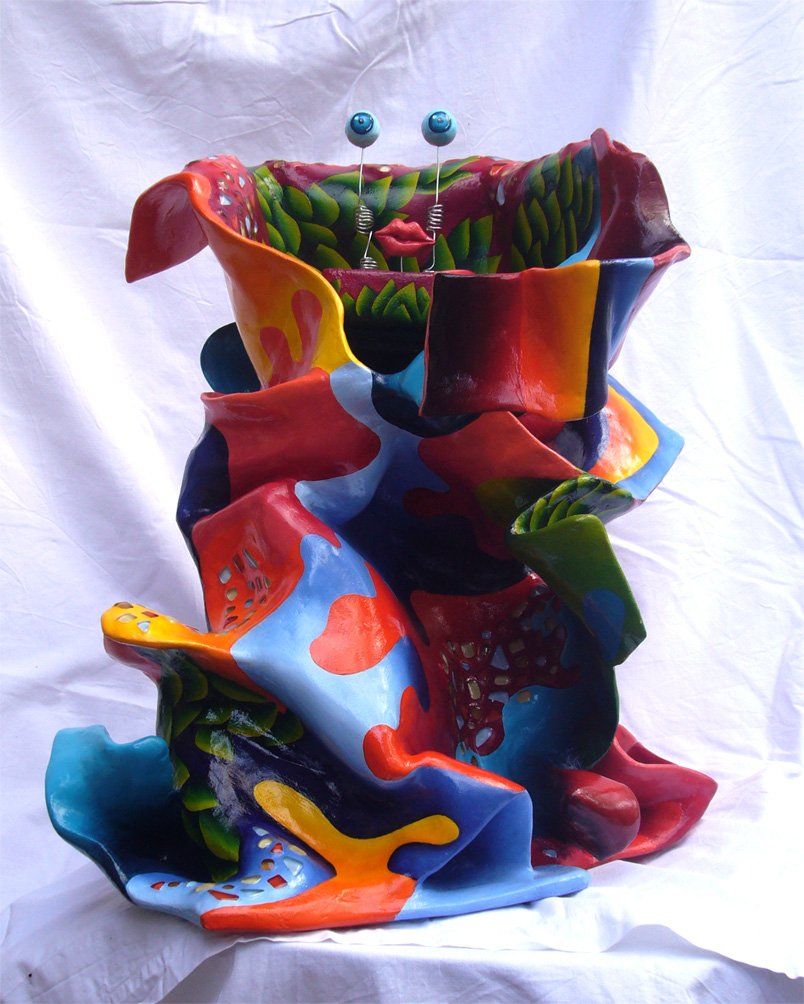 Abstrakte Plastik aus Pappmasché der Künstlerin Benita von Wendt. Titel: Elvis. Die abstrakte lebensfroh wirkende Skulptur ist sehr bunt mit Ornamenten die an Blätterwerk erinnern, sie scheint in einen Mantel mit sehr großem Kragen gehüllt. Im Innenraum des Kragens befindet sich eine Schublade, darauf sind mit Silberdraht zwei Augäpfel und ein Mund montiert.