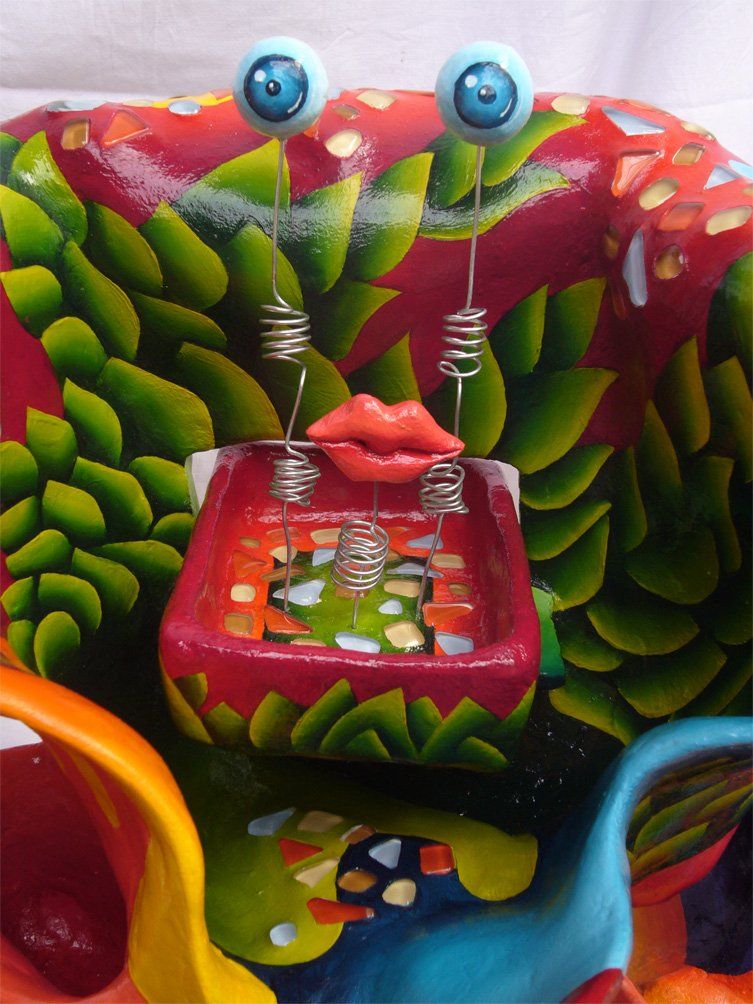 Abstrakte Plastik aus Pappmasché der Künstlerin Benita von Wendt. Titel: Elvis. Die abstrakte lebensfroh wirkende Skulptur ist sehr bunt mit Ornamenten die an Blätterwerk erinnern, sie scheint in einen Mantel mit sehr großem Kragen gehüllt. Im Innenraum des Kragens befindet sich eine Schublade, darauf sind mit Silberdraht zwei Augäpfel und ein Mund montiert. Detailansicht.