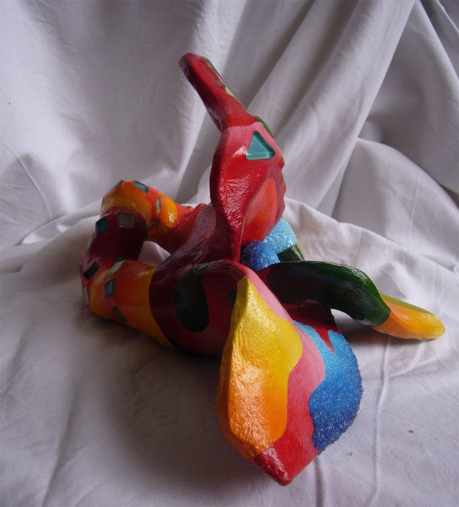 Plastik aus Pappmasché der Künstlerin Benita von Wendt. Titel: Fleur d'espoir 1. Bunte Skulptur die eine kleine vierblätterige Blume, die auf dem Boden liegt darstellt. Die Blume ist mit bunten, abstrakten Ornamenten bemalt und mit Mosaiksteinen besetzt. Seitenansicht.