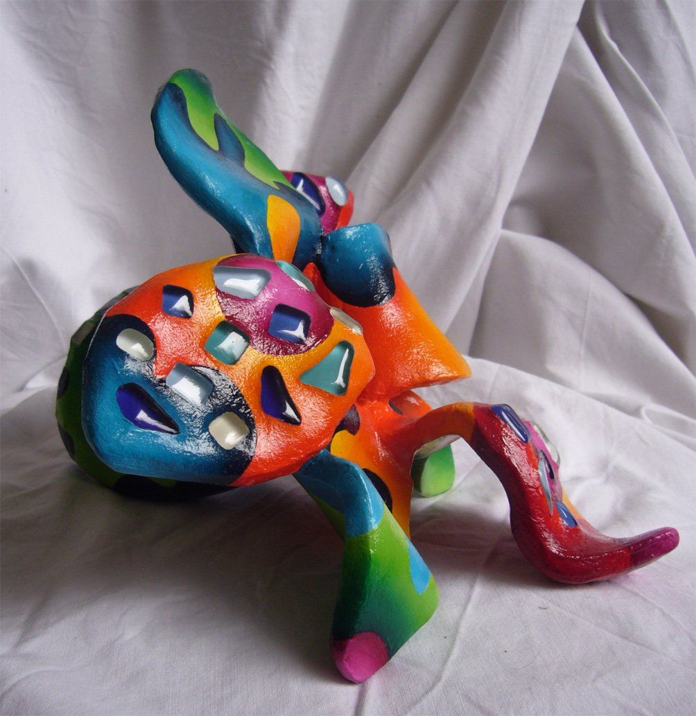 Plastik aus Pappmasché der Künstlerin Benita von Wendt. Titel: Fleur d'espoir 2. Bunte Skulptur die eine kleine sechsblätterige Blume, die auf dem Boden liegt darstellt. Die Blume ist mit bunten, abstrakten Ornamenten bemalt und mit Mosaiksteinen besetzt. Seitenansicht.