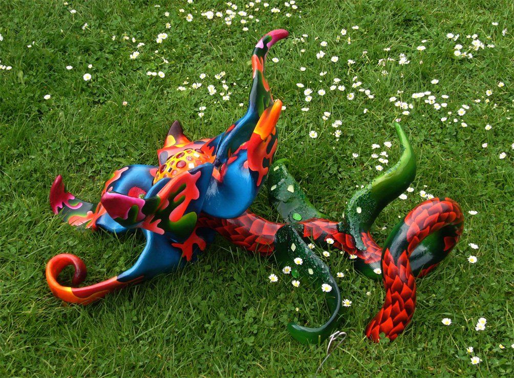 Plastik aus Pappmasché der Künstlerin Benita von Wendt. Titel: Blume im Gras. Bunte Skulptur die eine große Blume, die auf dem Boden liegt darstellt. Die Blume besteht aus einem Blütenstempel mit Mosaiksteinen besetzt, türkisen Blütenblättern mit abstrakten bunten Ornamenten, sowie einem grünen Stiel der mit Laub ähnlichen Formen bemalt ist. Seitenansicht halbrechts..