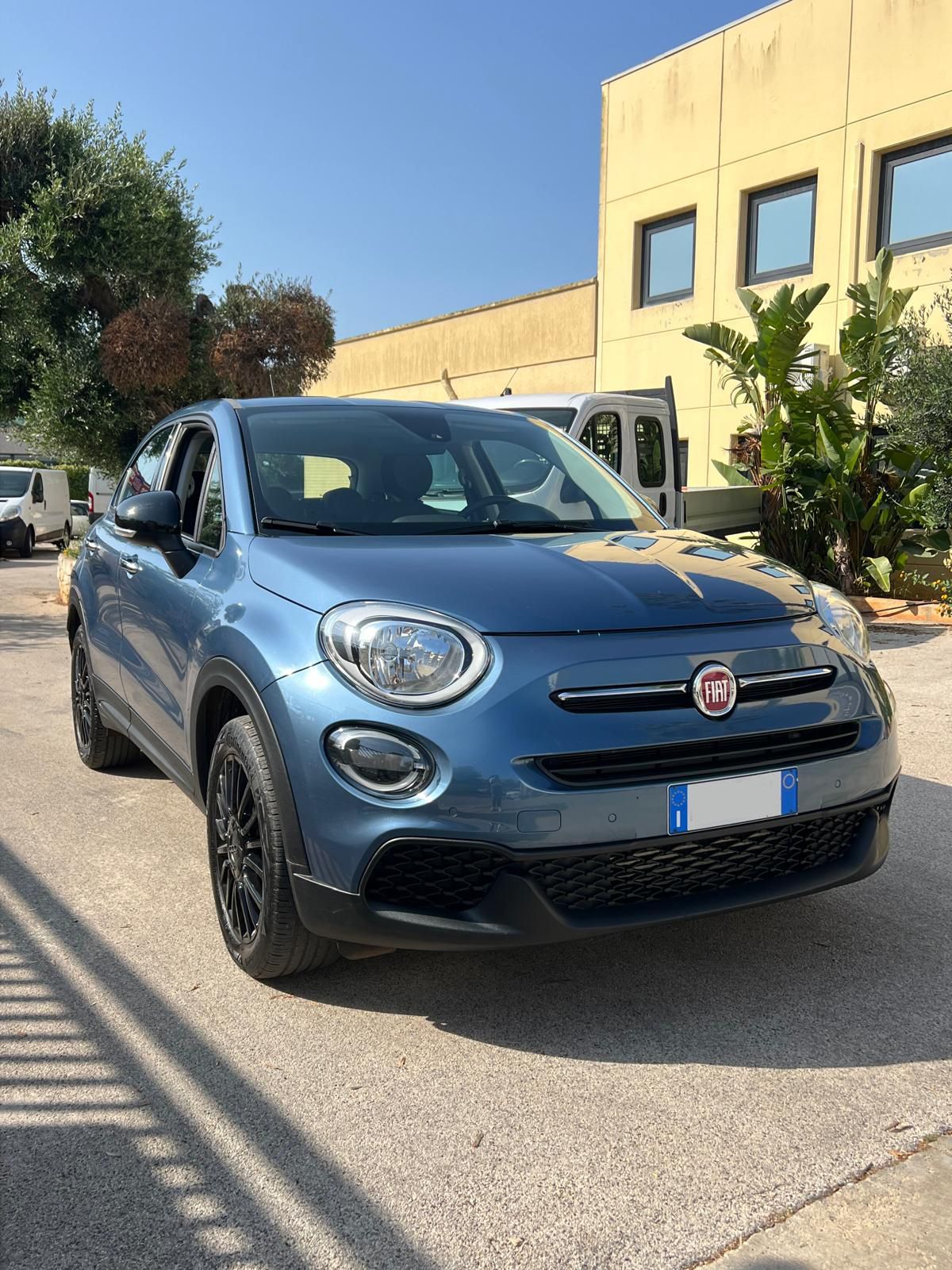 Un SUV Fiat 500X blu parcheggiato sull'asfalto di fronte a un edificio in una giornata di sole.