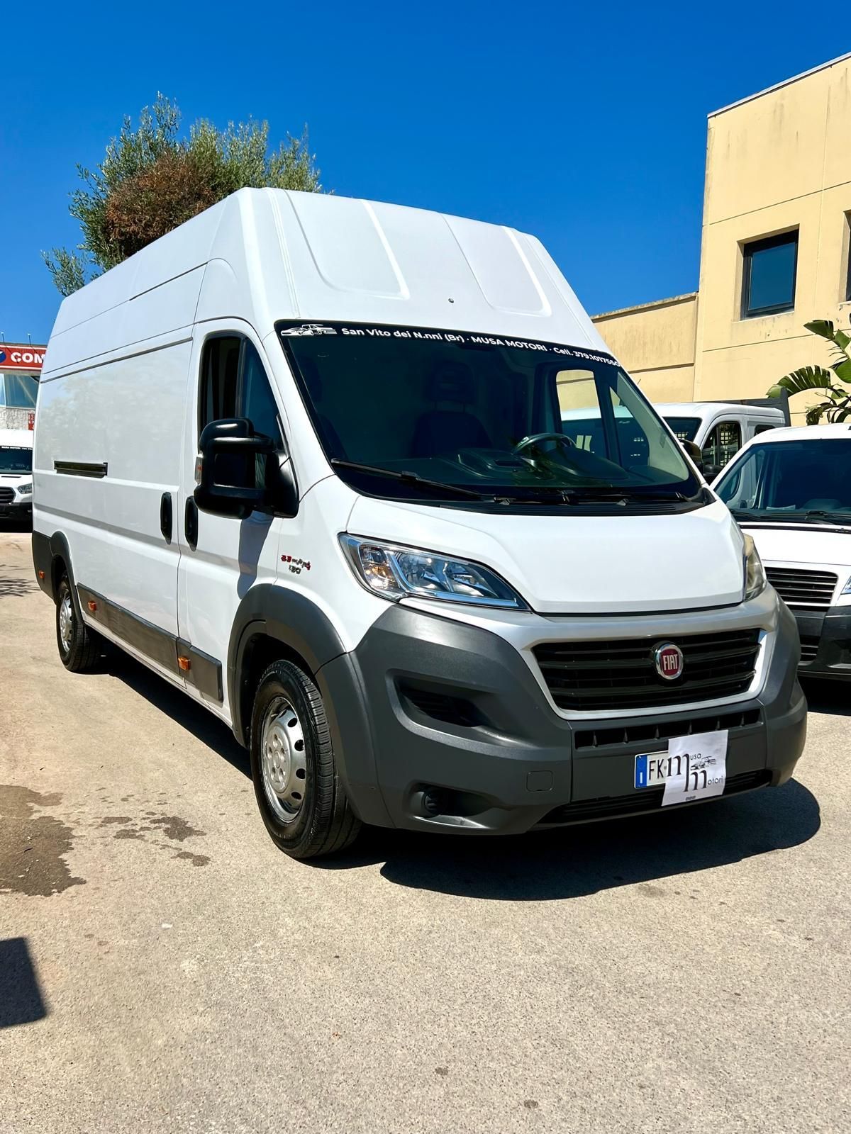 Furgone Fiat Ducato bianco, parcheggiato sul marciapiede, giornata di sole. Paraurti grigio, specchietti neri, sullo sfondo un edificio.