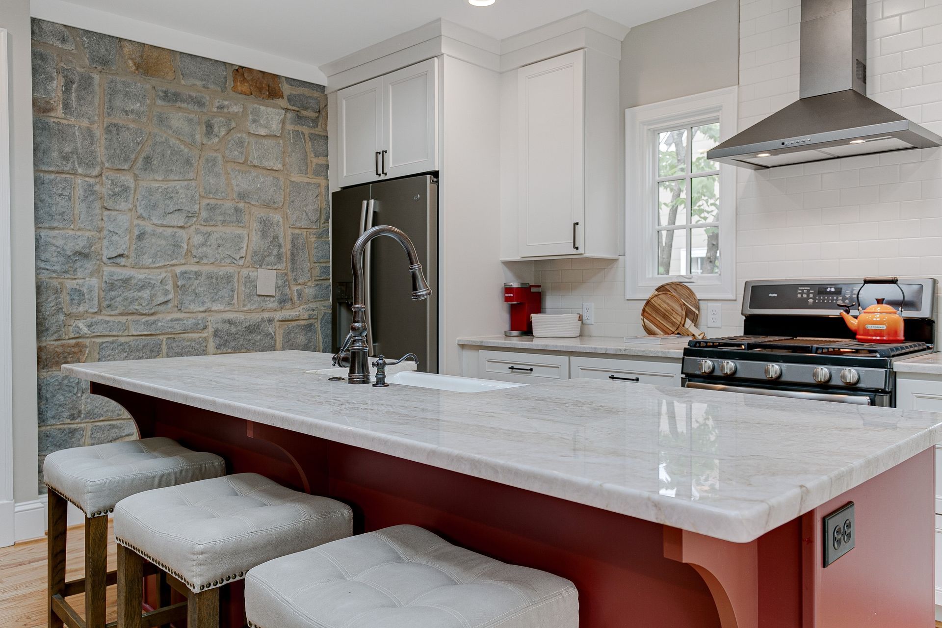 Kitchen with stone wall, white cabinets, red island, and gray stools.