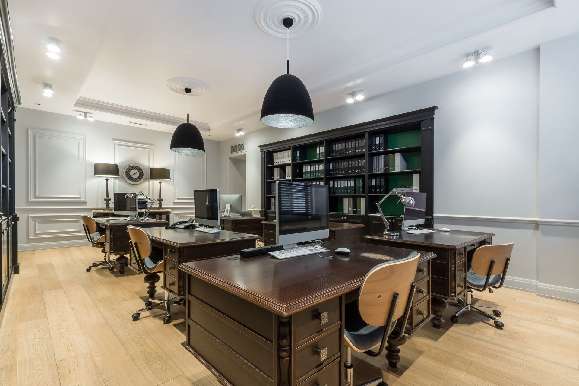 Office with wooden desks, chairs, and large bookcase; overhead black lamps; light wood floor.
