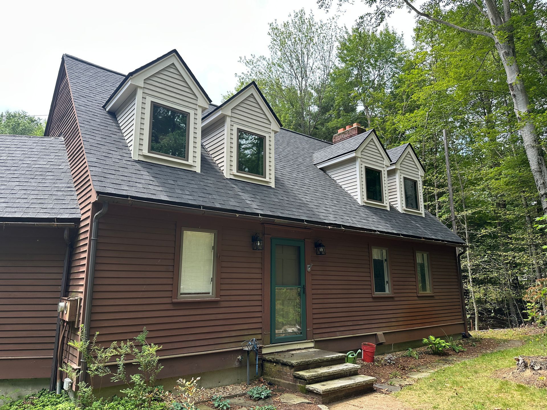 A brown house with a black roof and white trim is surrounded by trees.