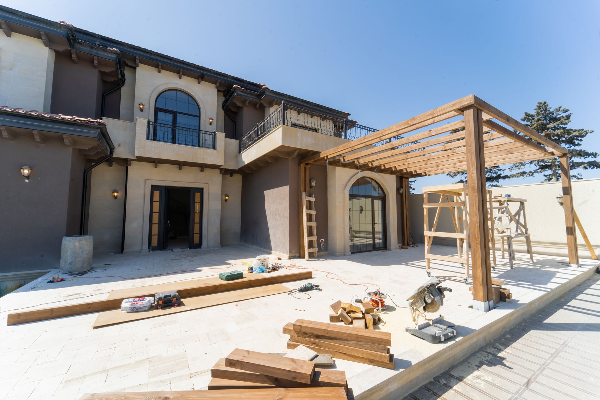 A house under construction with a wooden pergola. Construction tools and materials scattered on the concrete patio.