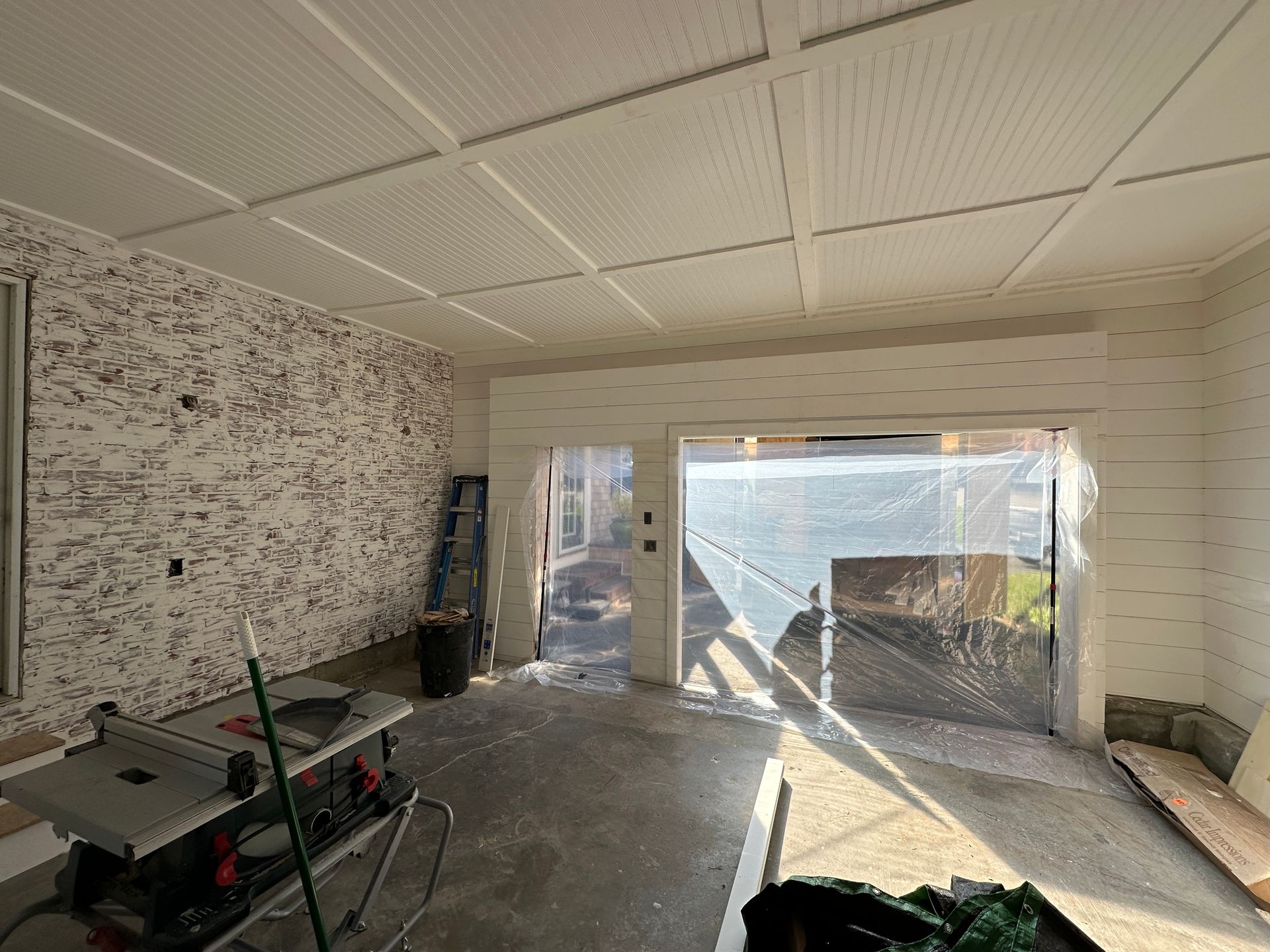 Empty room under renovation with unfinished walls, drop ceiling, and sunlight through large sliding glass doors.
