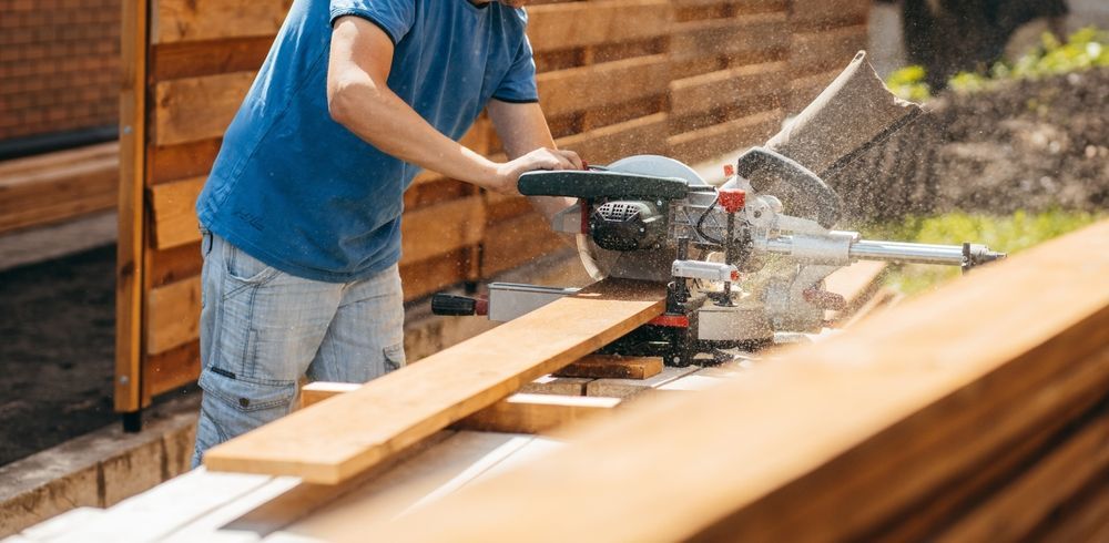 Person in blue shirt using a circular saw to cut a piece of wood outdoors, near a wooden fence.