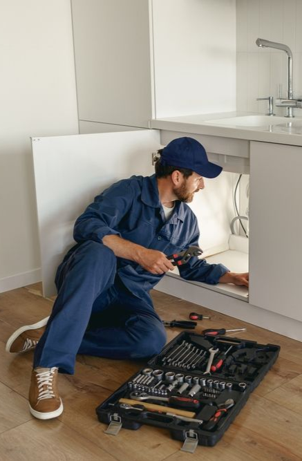 Plumber in blue jumpsuit, cap, examining pipes under a sink, holding wrench, tool kit nearby.