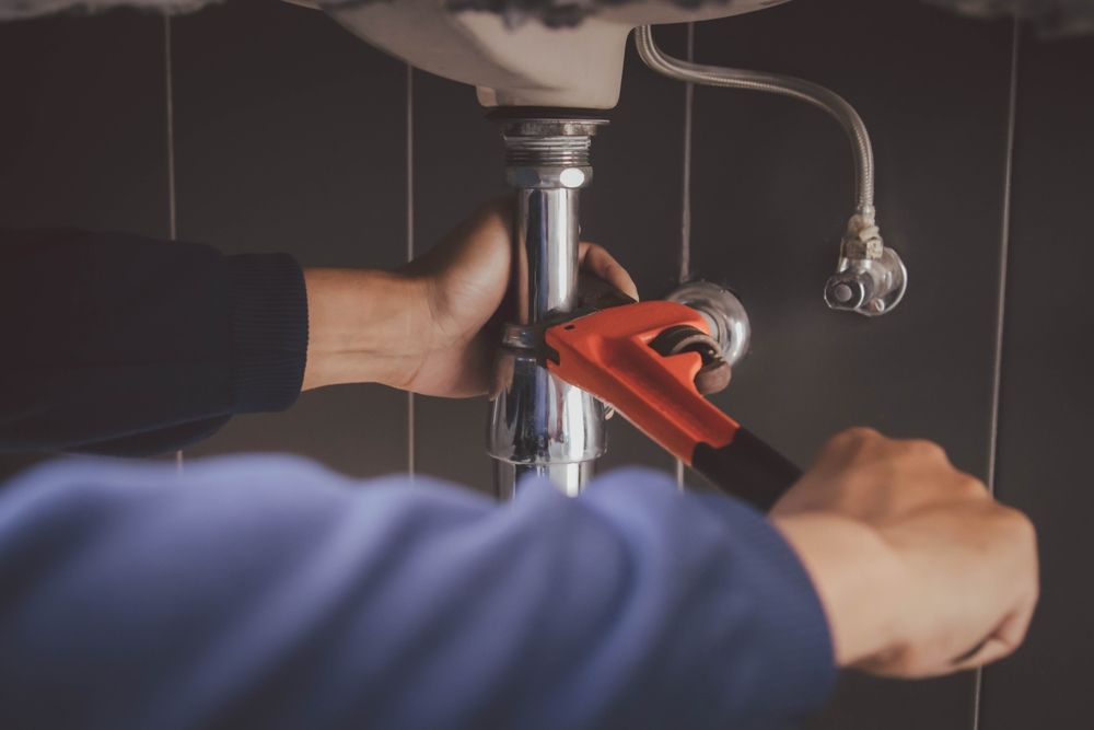 Plumber tightening a pipe under a sink with an orange wrench in a dark bathroom.