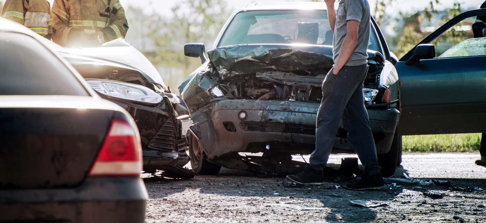 A car accident scene. Three damaged vehicles. A person stands near a car, others are possibly first responders.