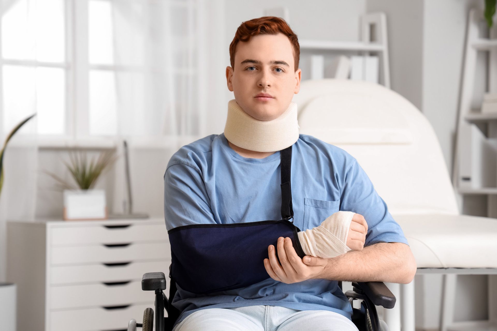 Man in wheelchair with neck brace and arm sling, in doctor's office setting.