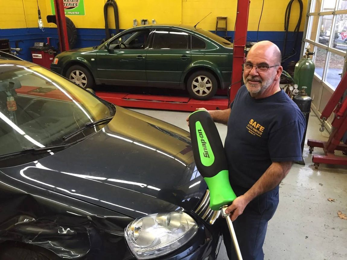 Man holding a green and black tool in a garage, standing by a black car and a green car.
