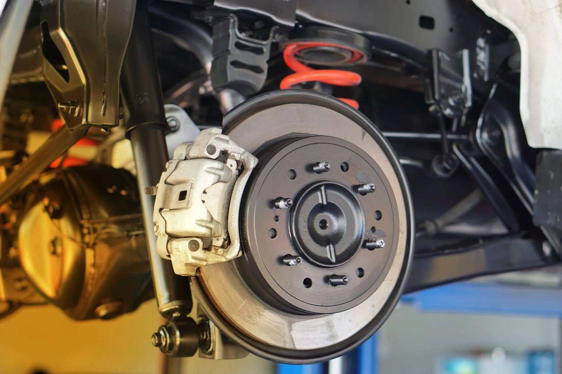 Rear car wheel with brake components visible; silver caliper, brake rotor, and red spring.
