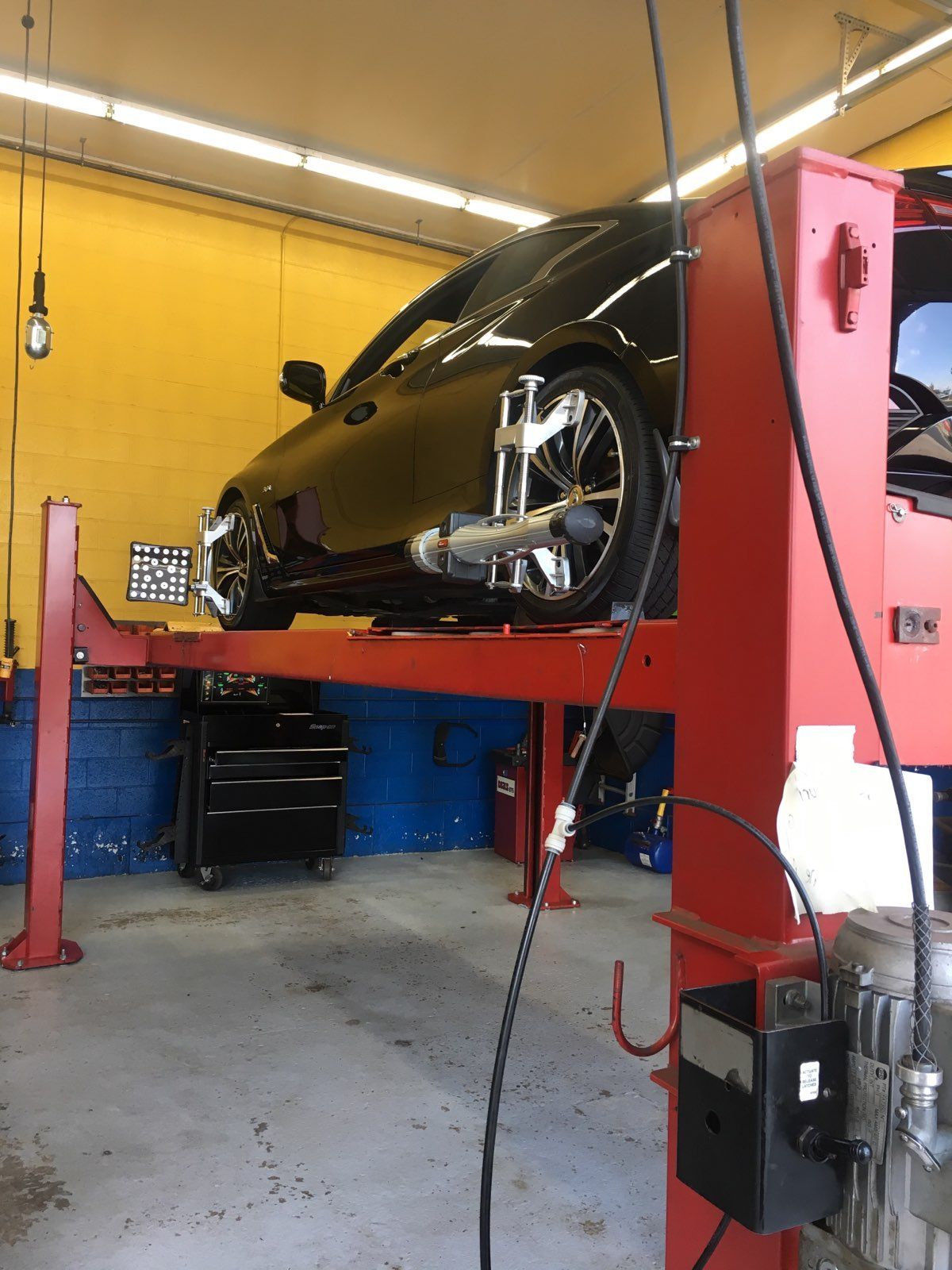 Black car on a red lift, undergoing wheel alignment in a repair shop.