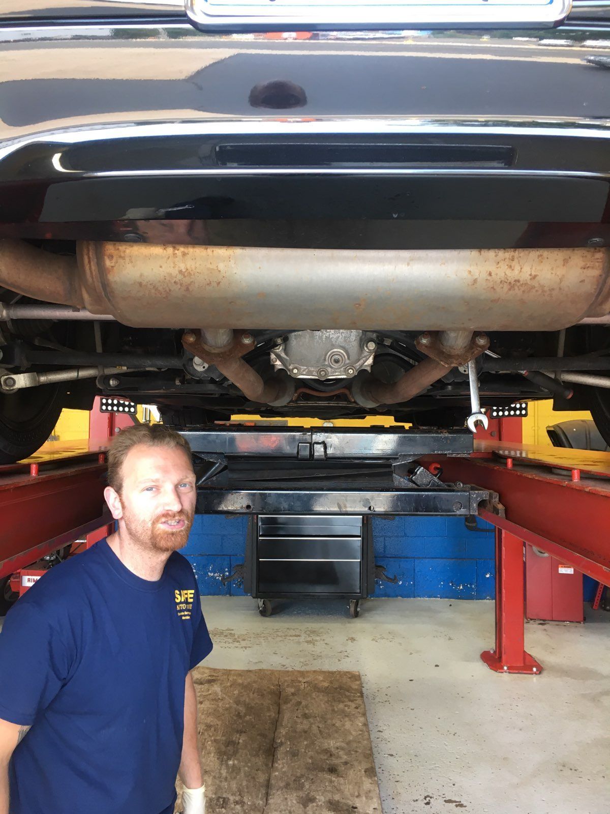 Mechanic under car on a lift; looking at camera. Garage setting, dark car, rusty muffler, blue shirt.