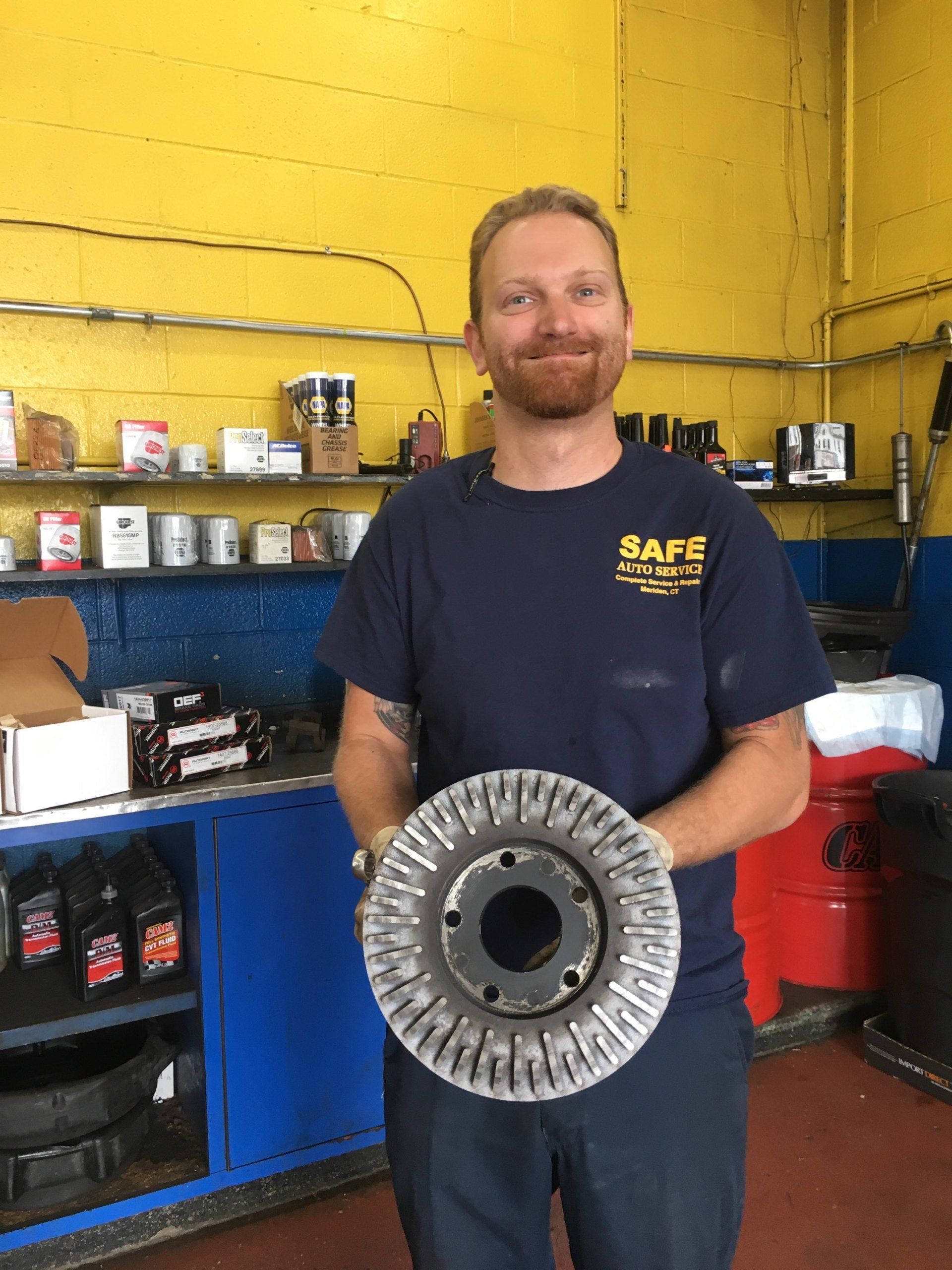 Mechanic holding a large metal part, smiling in a garage with blue cabinets and yellow walls.