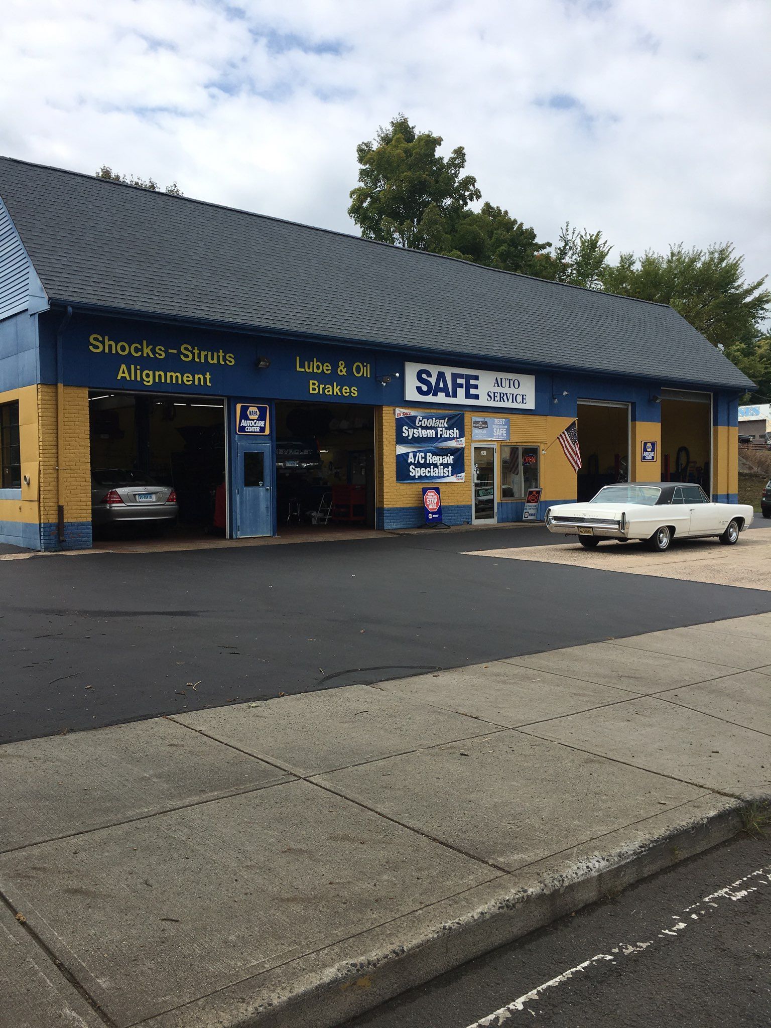 Auto repair shop with a blue and yellow facade; a white car sits inside and a classic white car is parked outside.