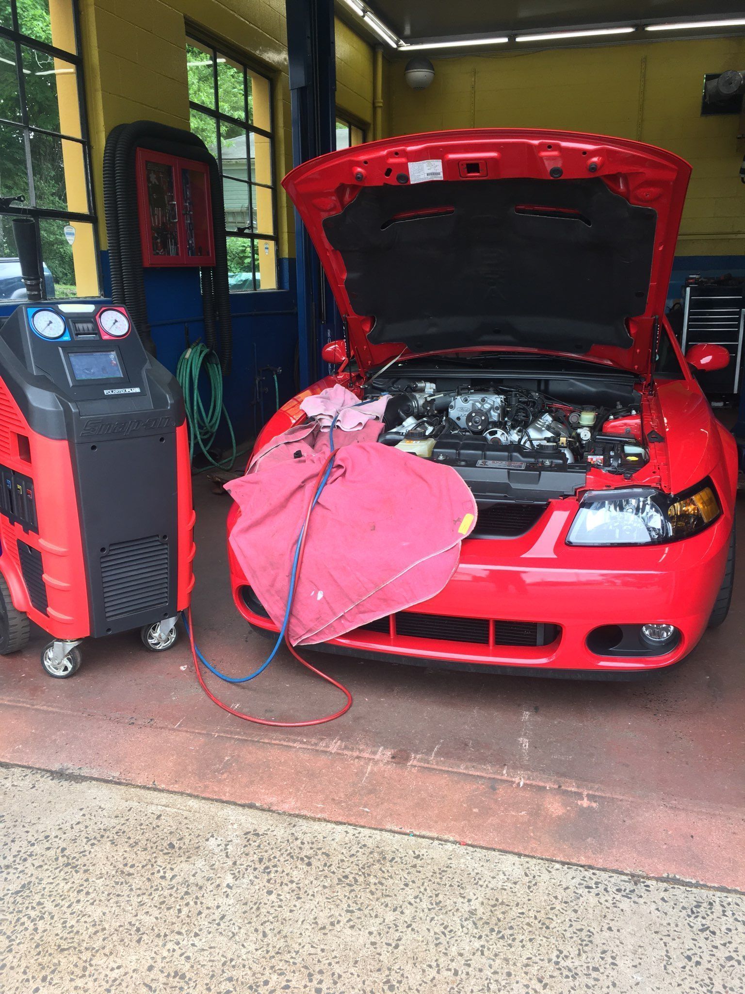 Red car with hood open being serviced in a garage with an A/C machine attached.
