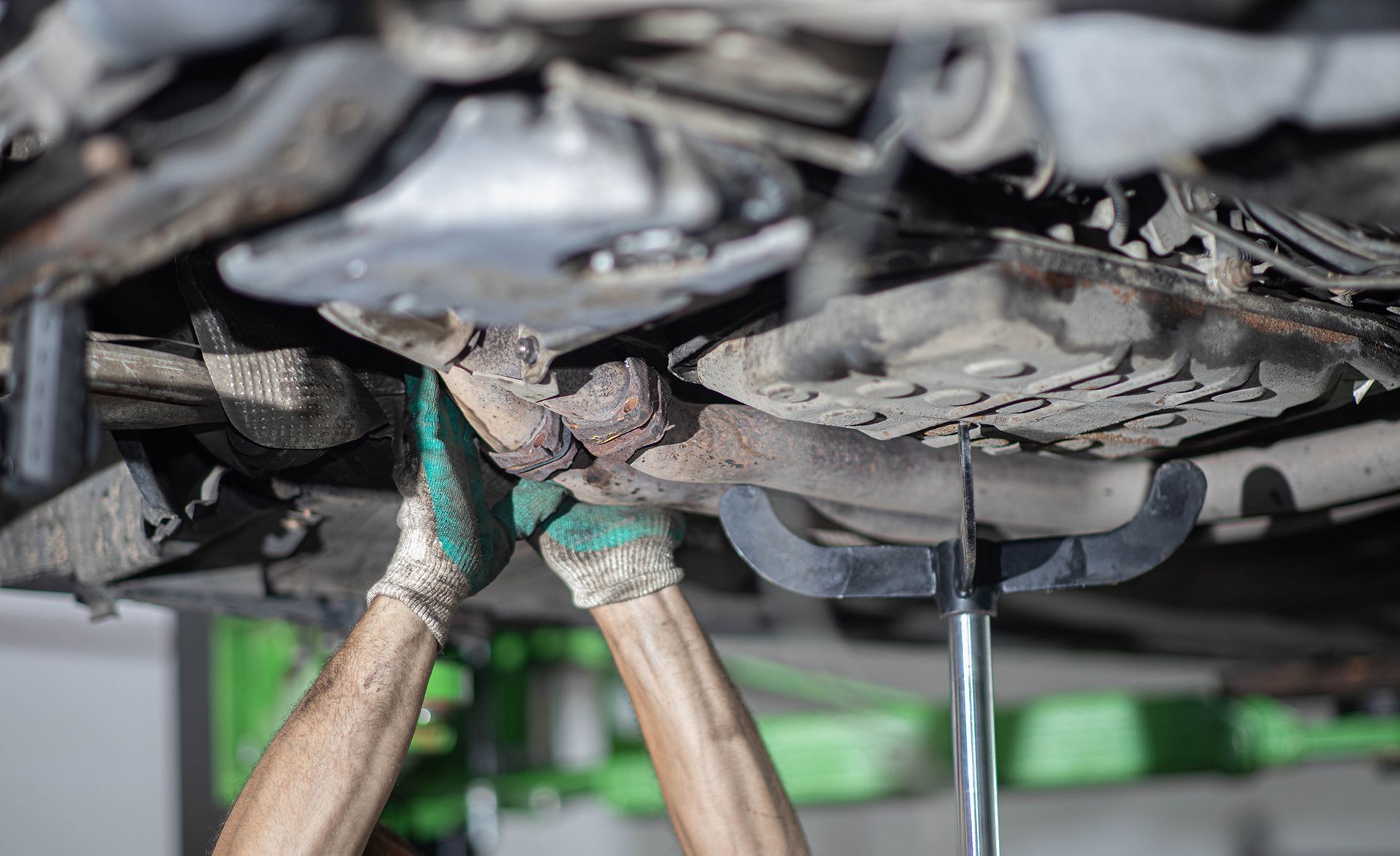 Hands in work gloves hold up a car exhaust pipe while working under a vehicle.