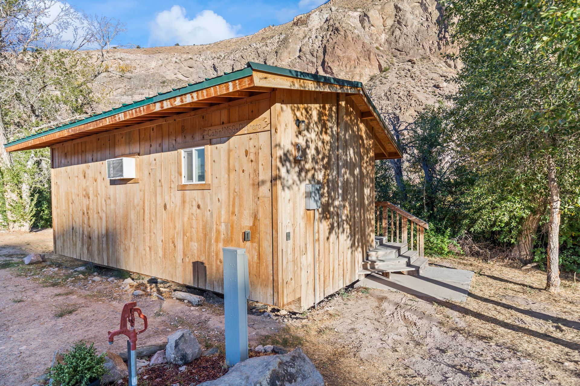 Wooden cabin with green roof, next to concrete path and faucet, set against a rocky mountain.
