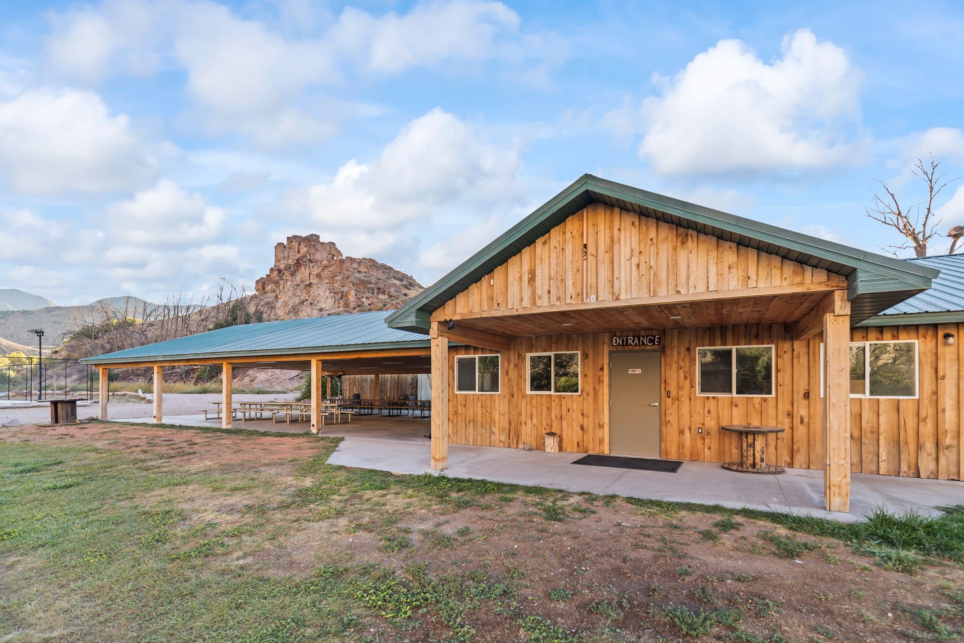 Wooden building with a covered patio, a mountain backdrop, and a grassy foreground under a cloudy sky.