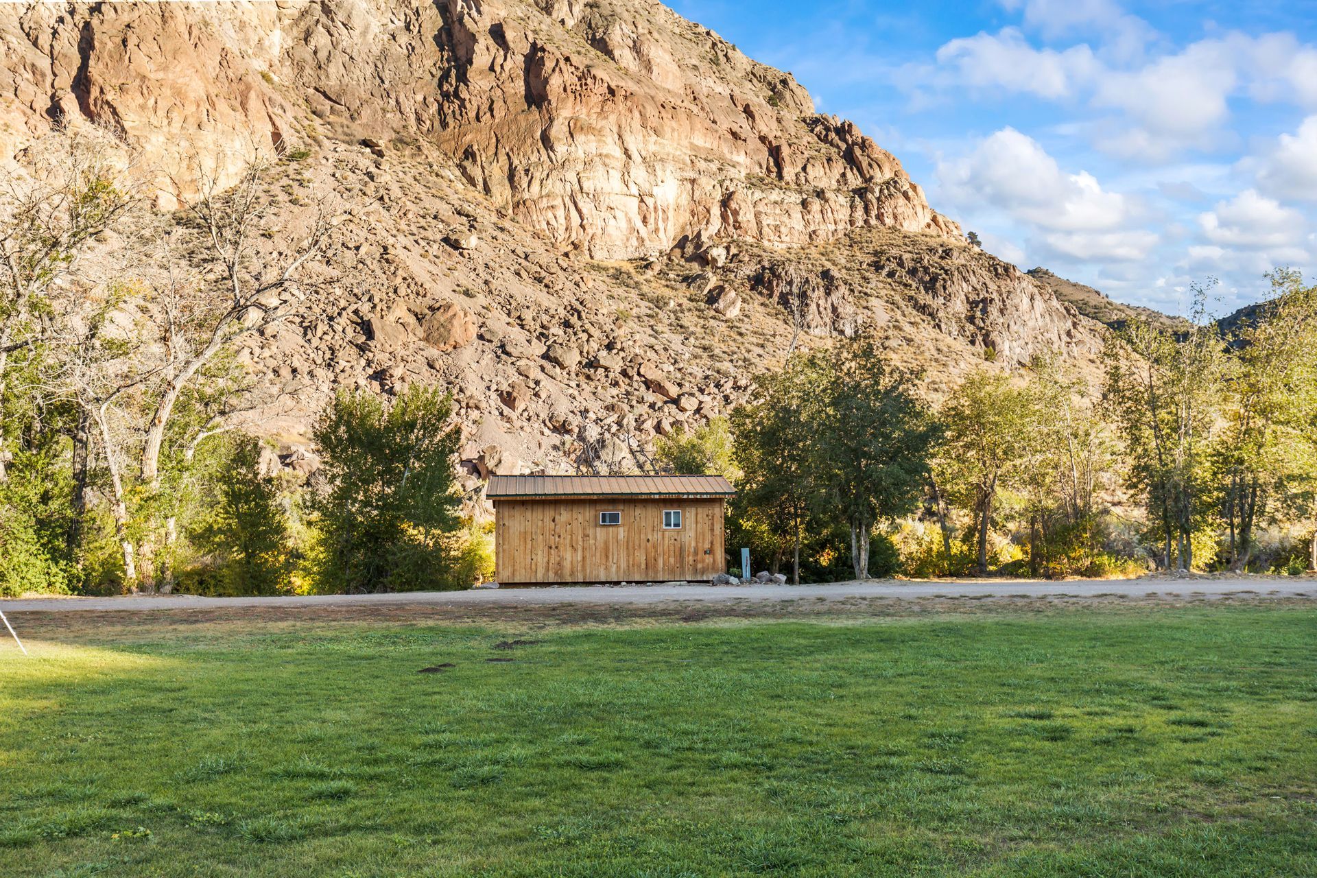 Wooden cabin in a grassy field, with a mountain backdrop and trees.