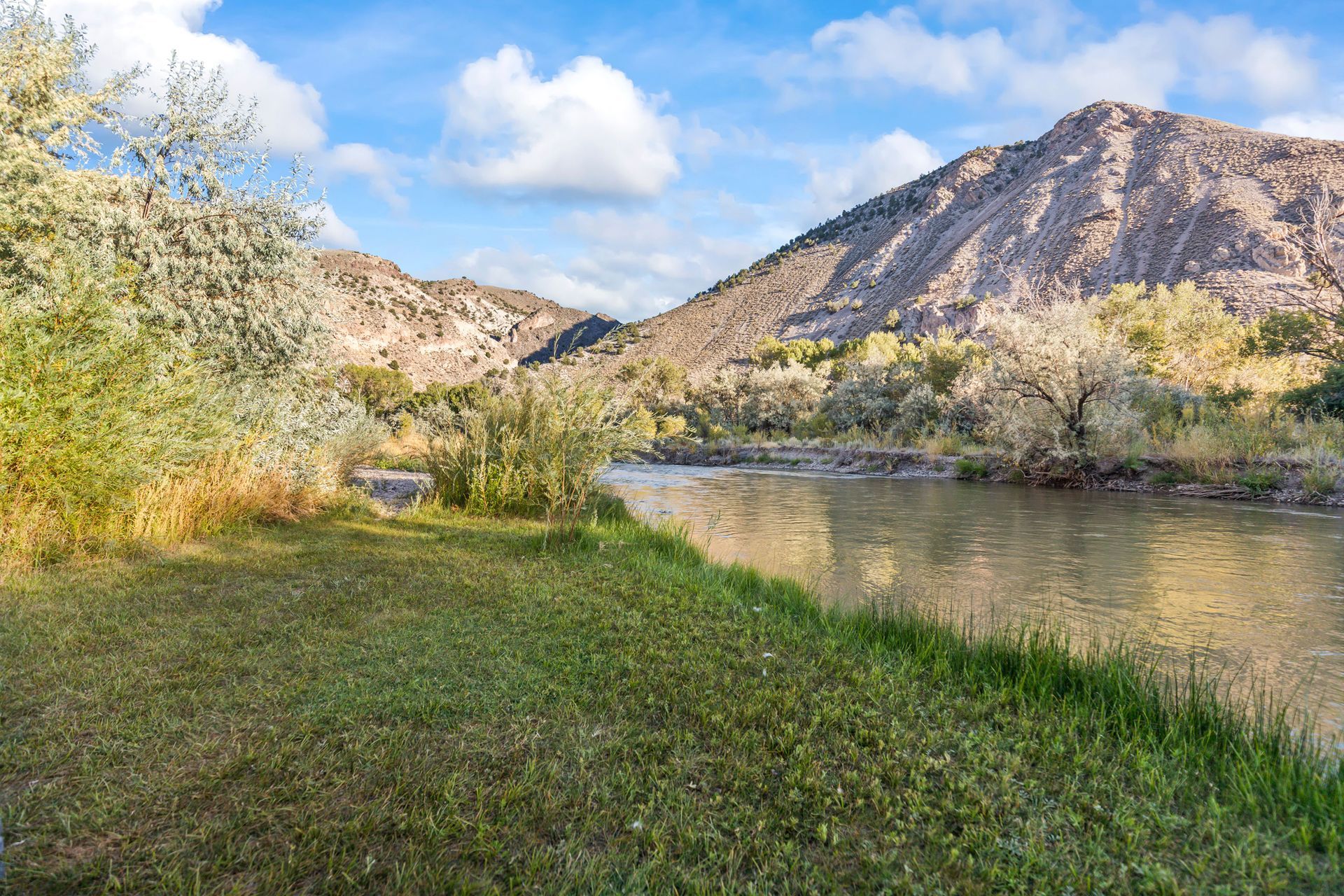 River flowing through a grassy bank, with mountains in the background under a blue sky with fluffy clouds.