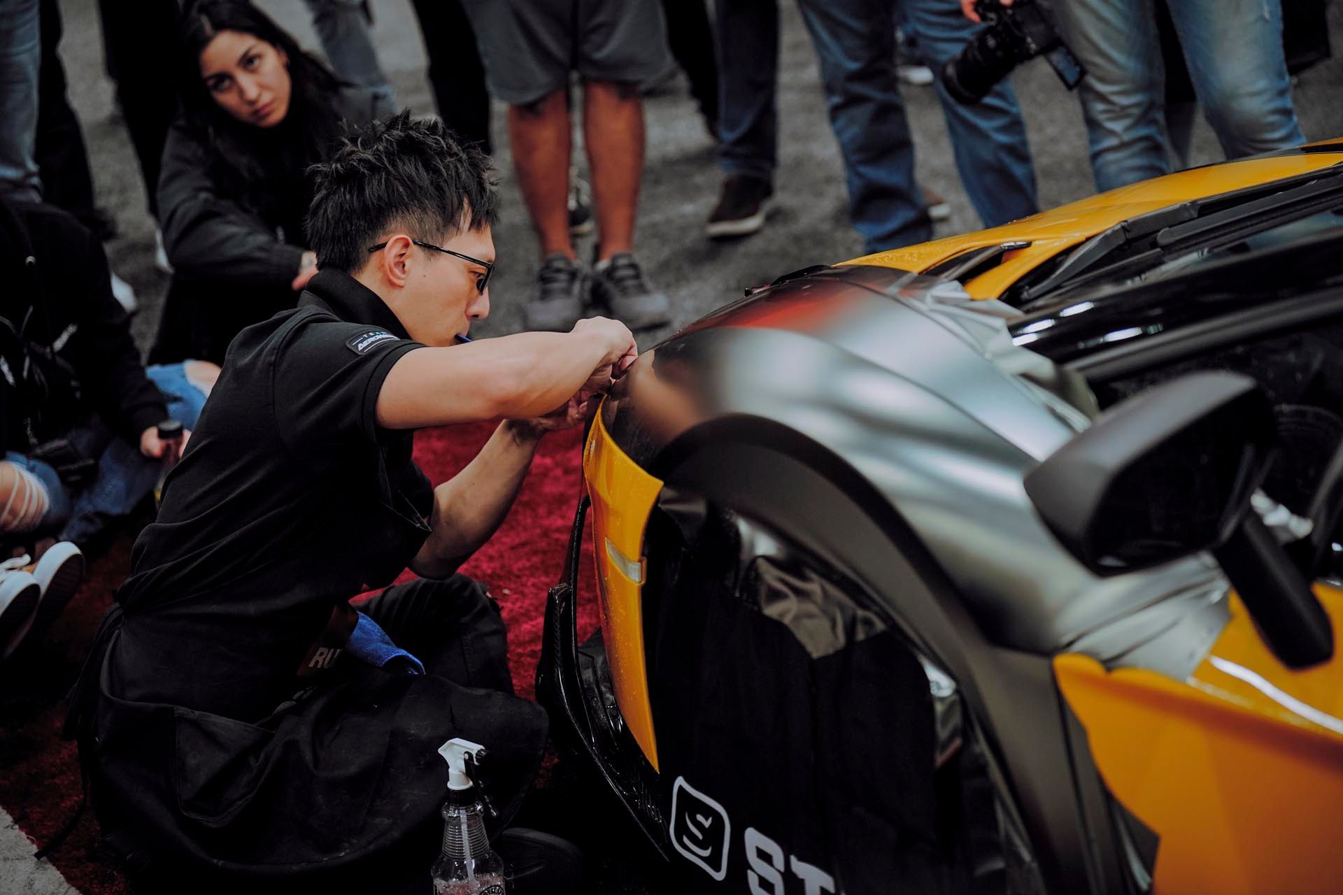 Man applying wrap to a yellow sports car; onlookers, indoor setting.