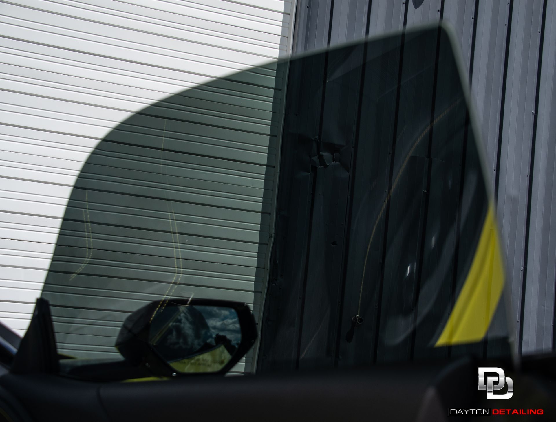Car window with tint, reflecting a wheel and yellow accent against a wood siding.