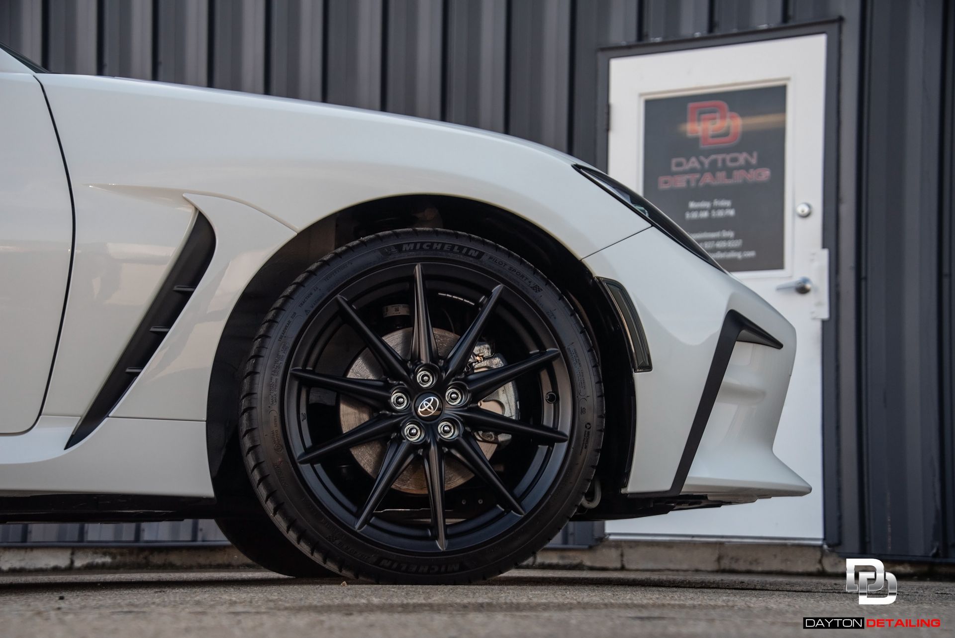 White sports car's front wheel, black rim, parked outside a Dayton Detailing shop.