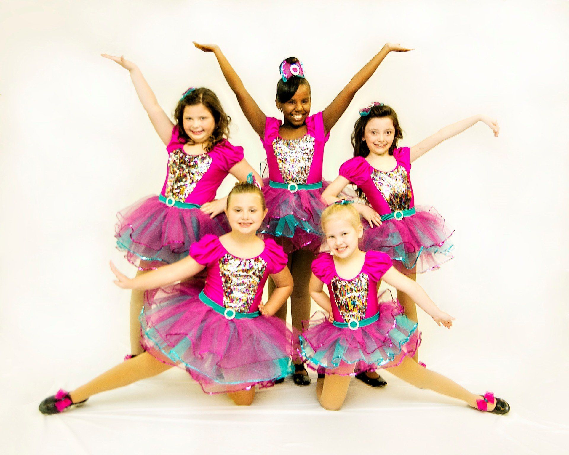 Group of young dancers in pink and blue tutus, posing with arms raised, smiles.