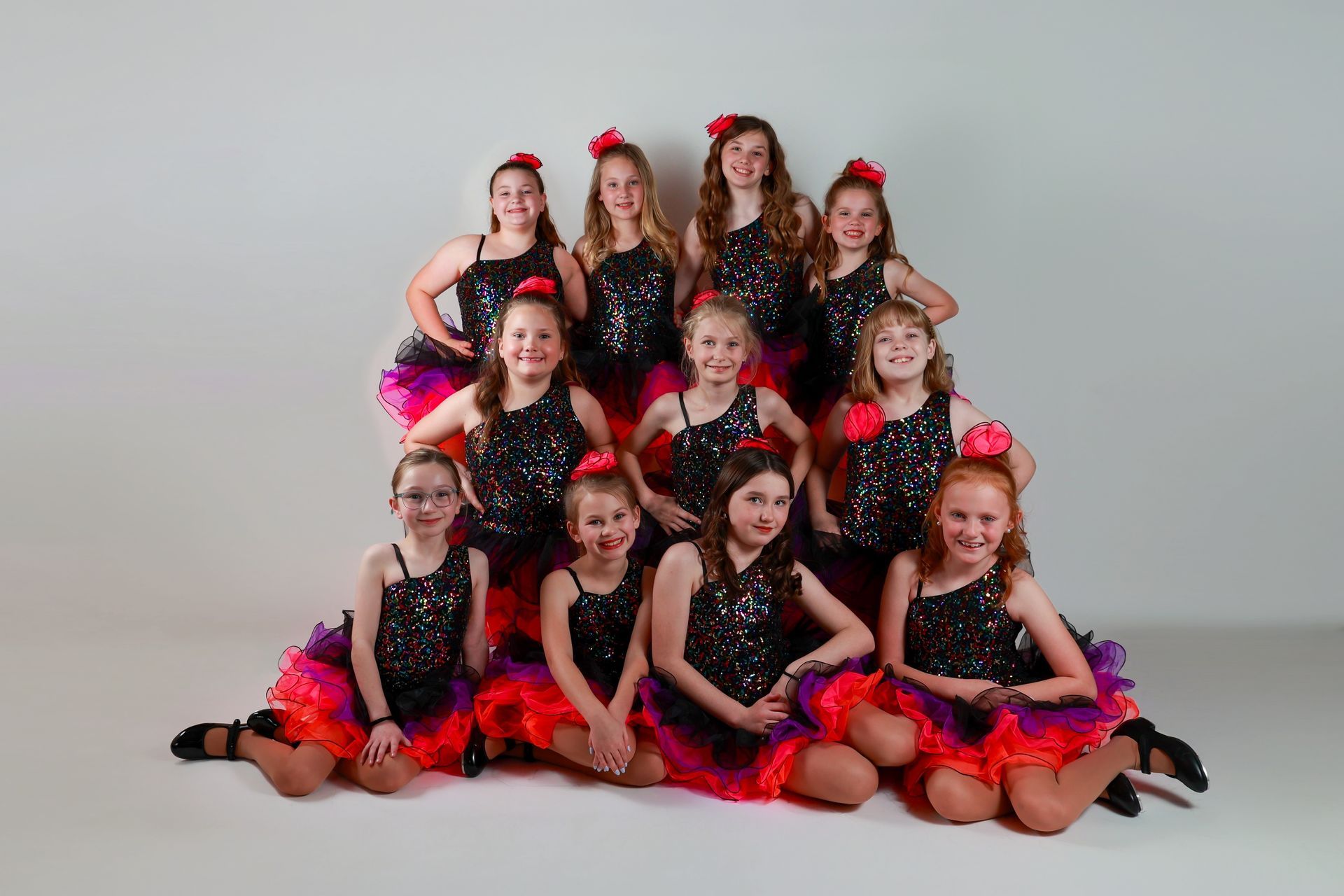 Group of young dancers in black sequin tops, colorful tutus, and red flower hair clips pose together.