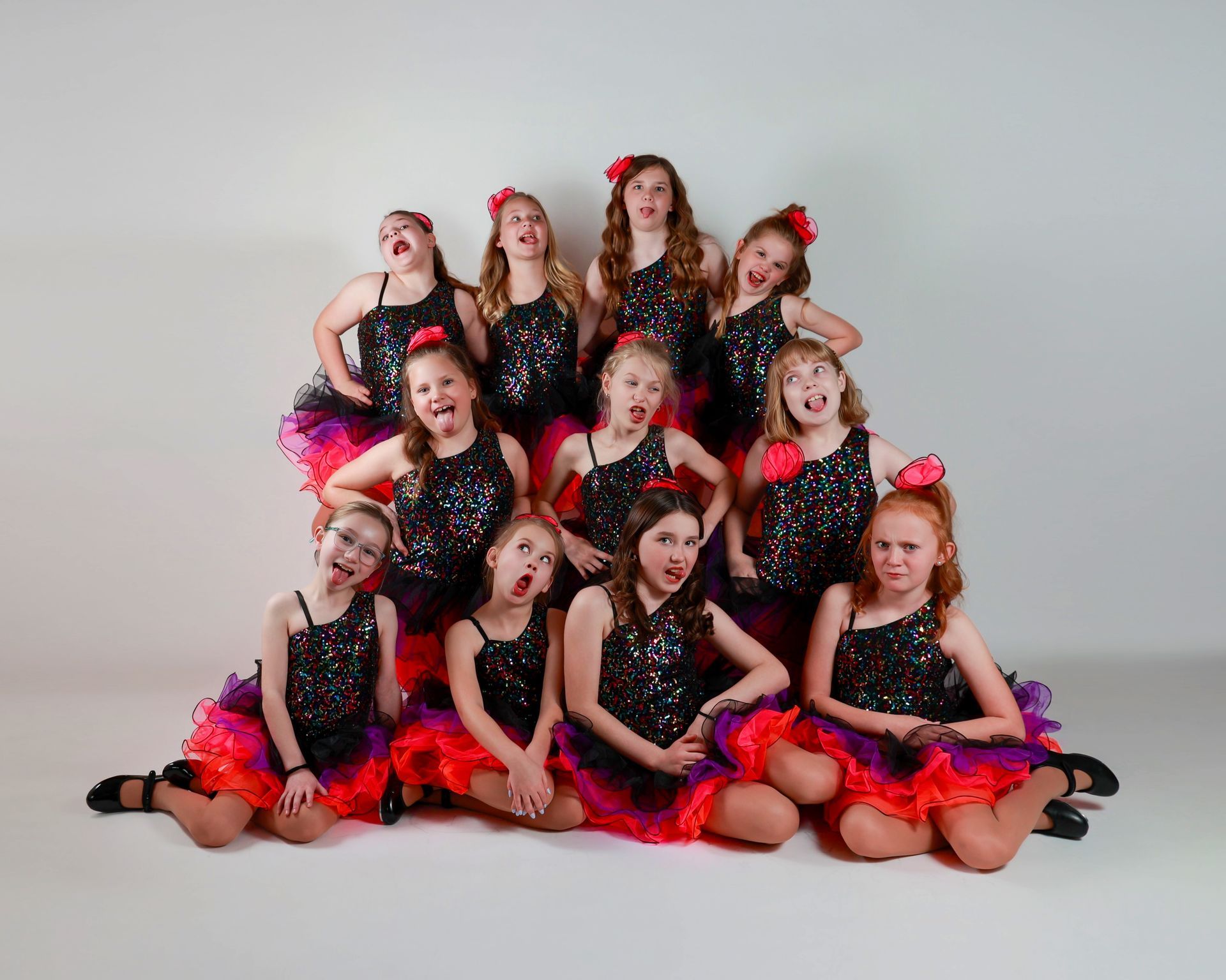 A group of young dancers in sparkly black tops and layered, colorful skirts make funny faces.