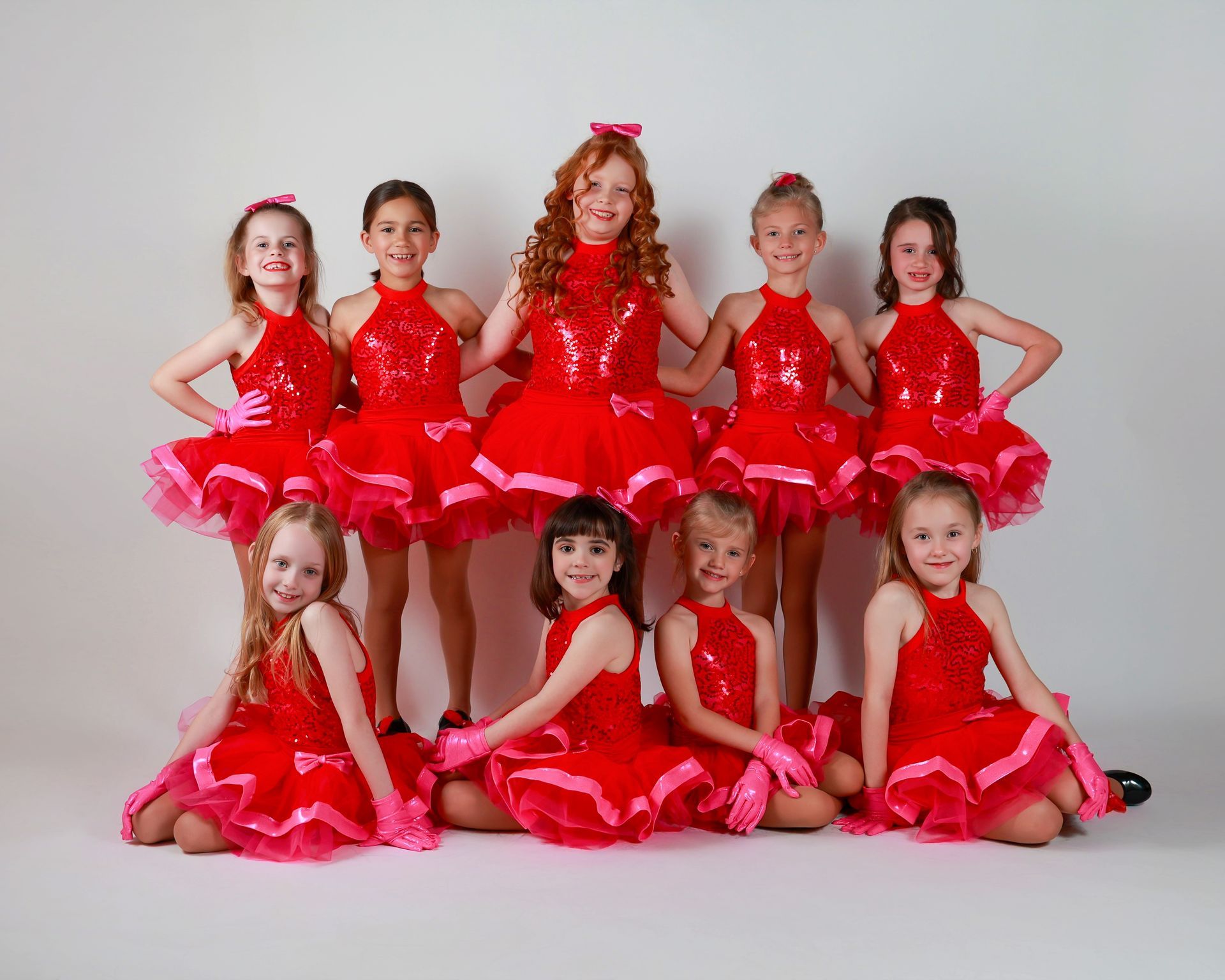 Nine young girls in red dance costumes, posing for a group photo on a white background.