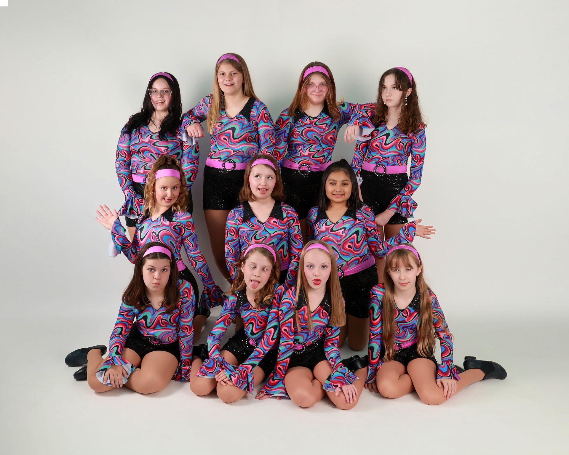 Dance team in colorful patterned tops, black shorts, and headbands, posing against a white backdrop.
