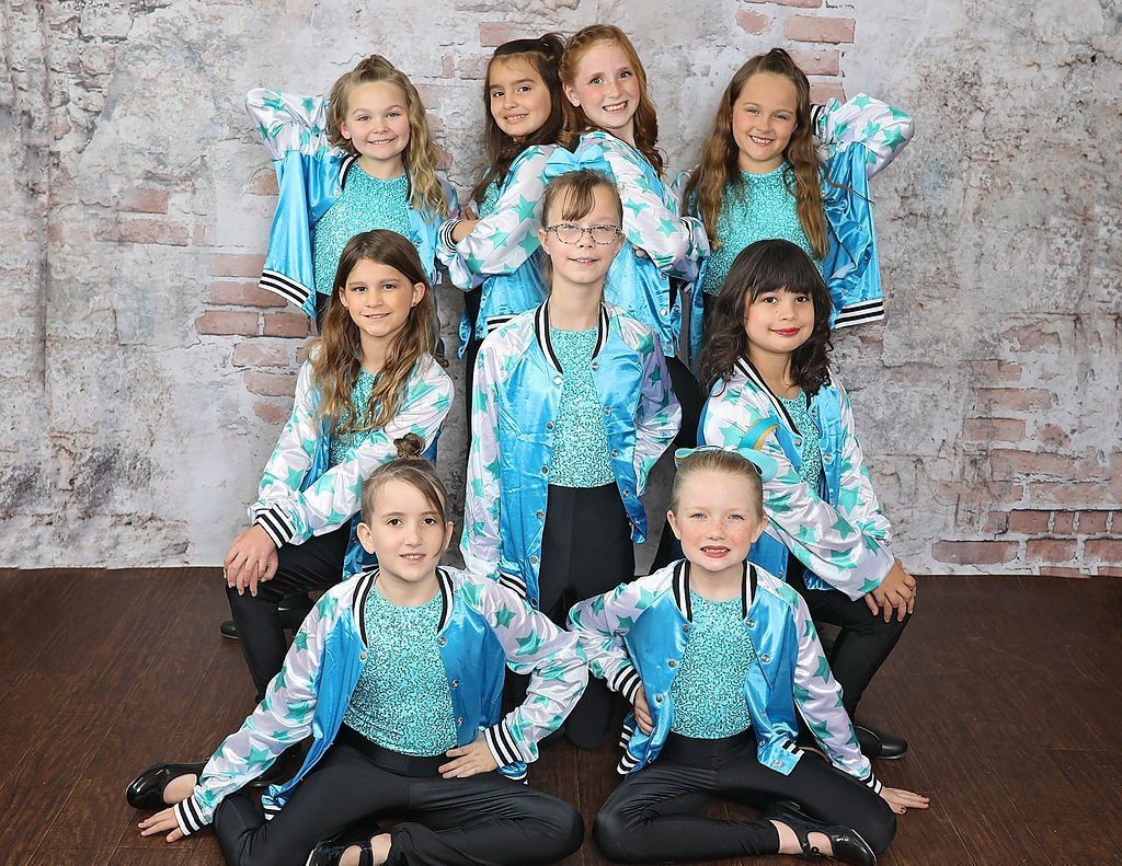 Group of young girls in turquoise jackets and black pants posing against a brick wall.