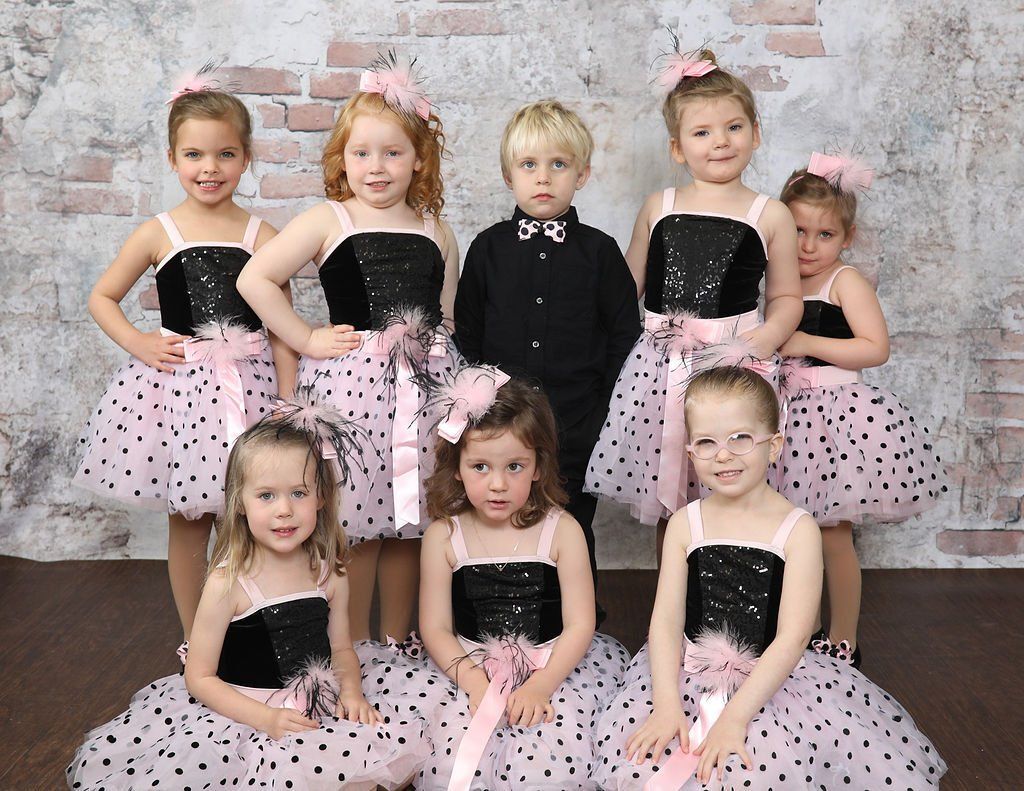 Group of young dancers in black and pink dresses, posing in front of a brick wall background.