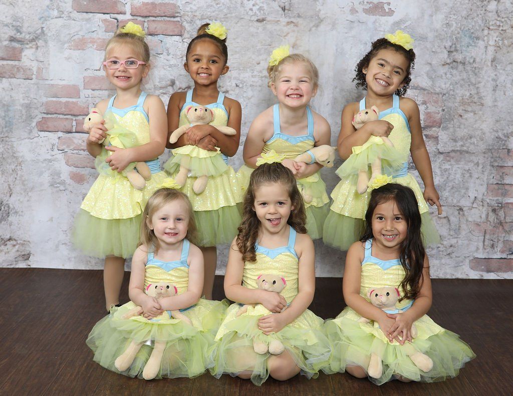 Group of young girls in yellow tutus, holding stuffed animals, posing for a photo. Brick background.