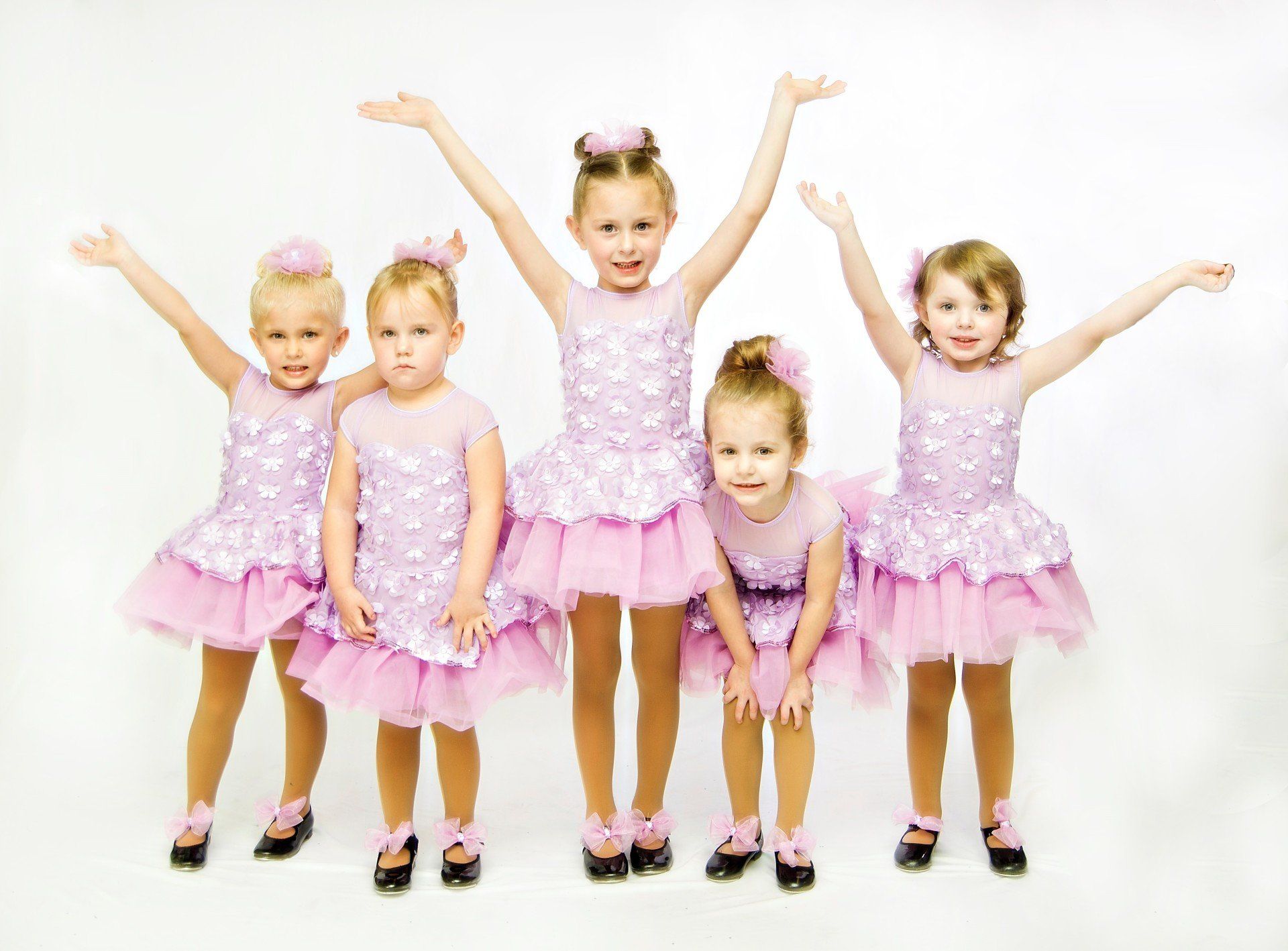 Five young girls in lavender dance costumes pose, arms raised, against a white background.
