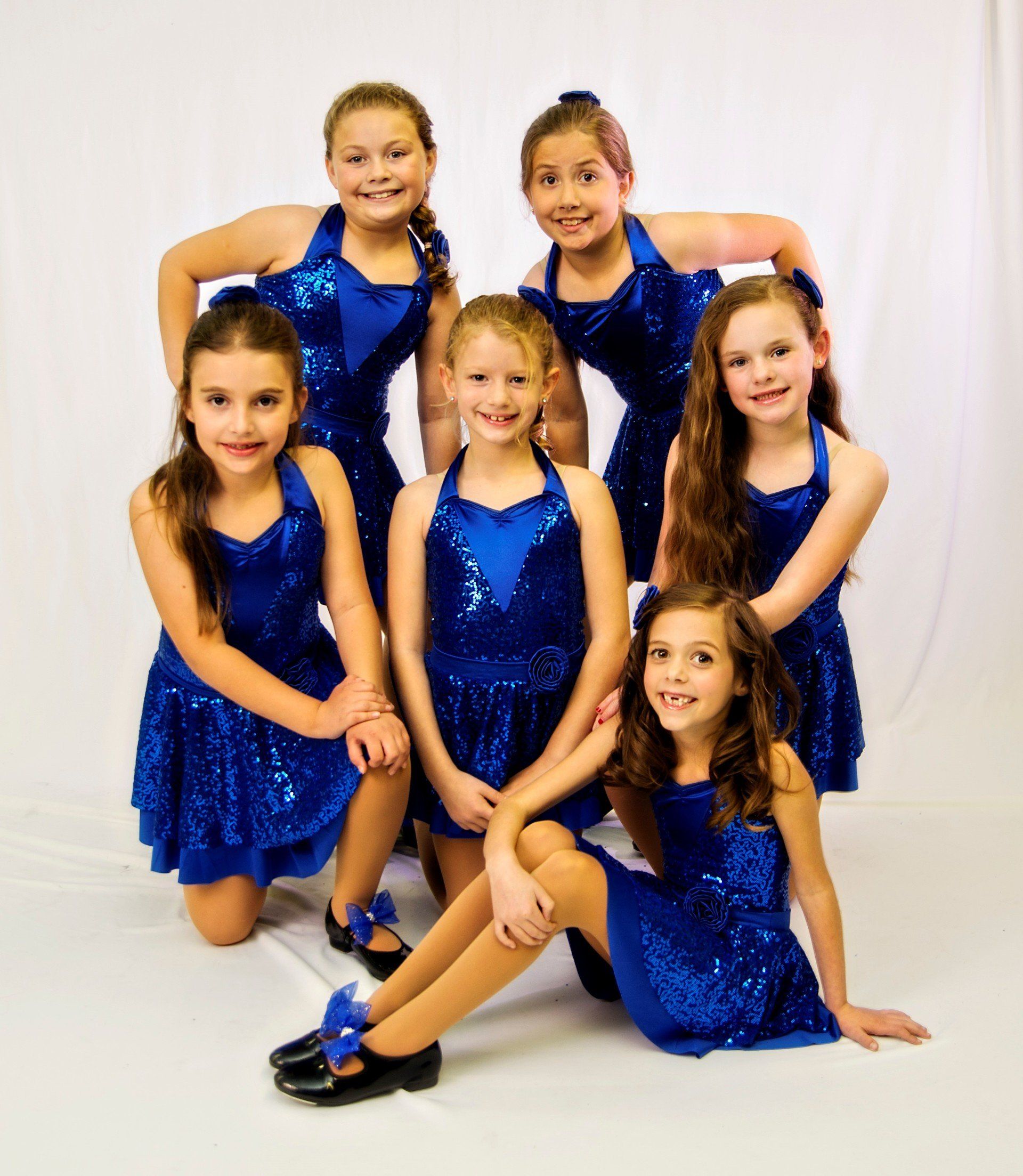 Six young girls in matching blue sequin dresses pose for a photo, smiling in a studio setting.