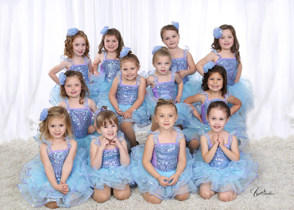 Group of young girls in blue dresses, posing for a photo. They are smiling and have bows in their hair.