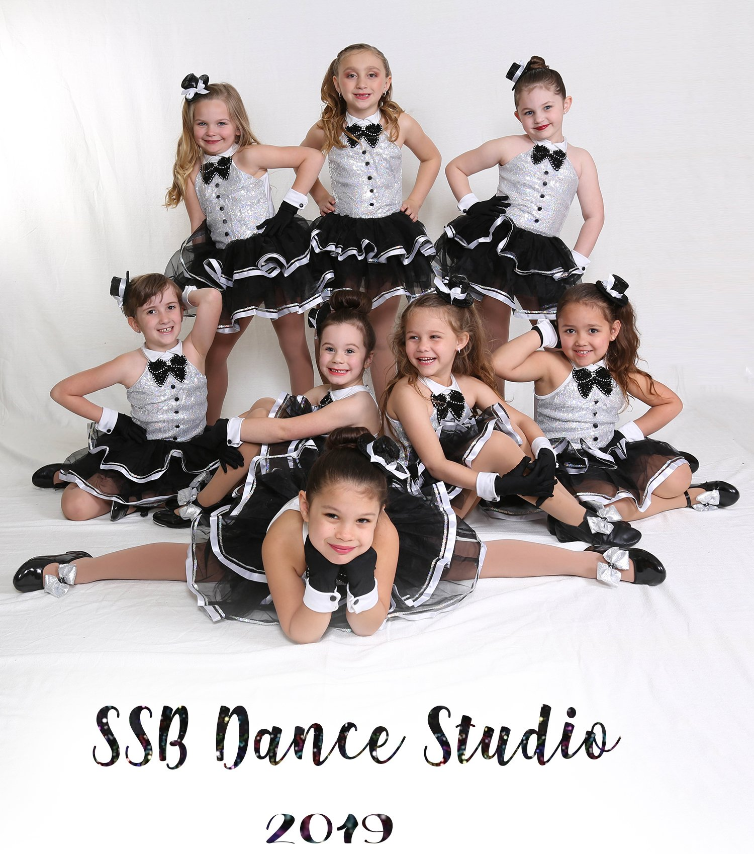 Nine young dancers in silver tops, black skirts, posing for a studio photo.