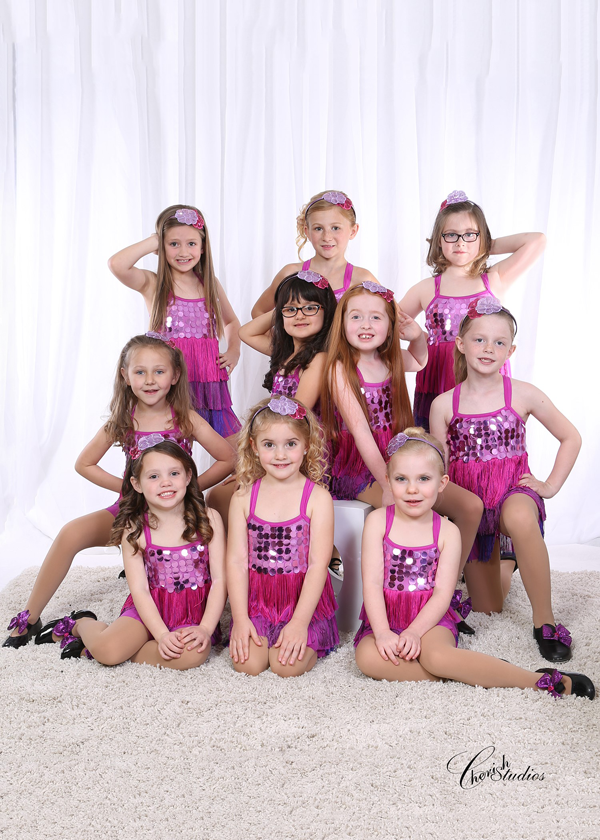 A group of young girls in pink sequin dance costumes, posing on a white rug.